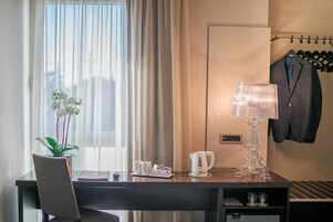 Habitación de hotel con escritorio, orquídea en maceta, lámpara, tetera y perchero con un traje.