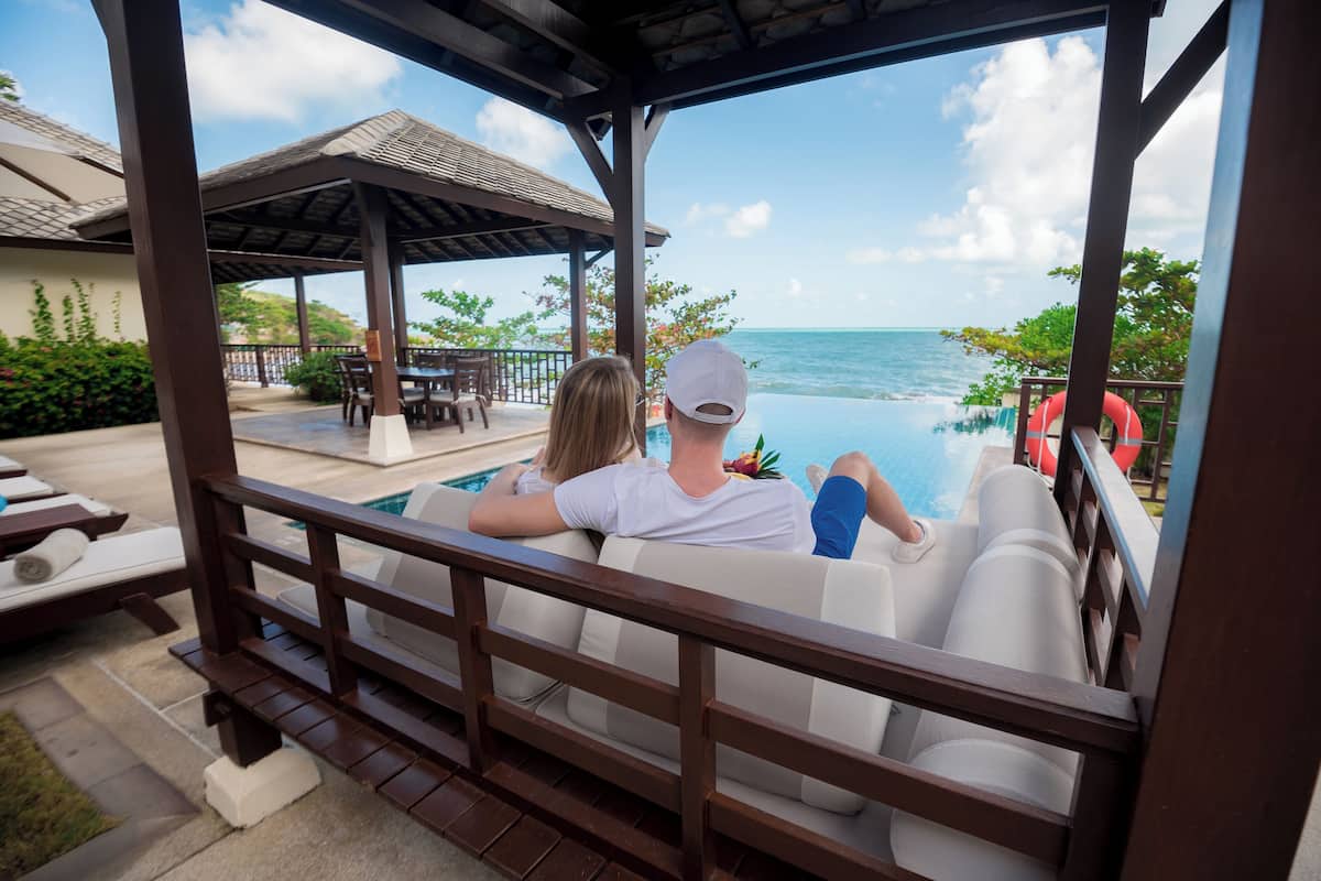 A couple relaxing by an infinity pool with a view of the ocean, wooden deck, and lounge chairs.