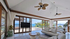 A spacious living room with a large window overlooking the sea, a sofa, a coffee table, and ceiling fans. - Kanda Residences Pool Villas (Koh Samui)