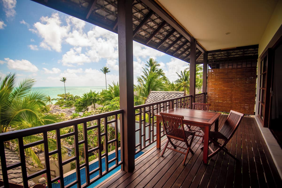 A wooden deck with a table and chairs, overlooking a pool and tropical scenery.
