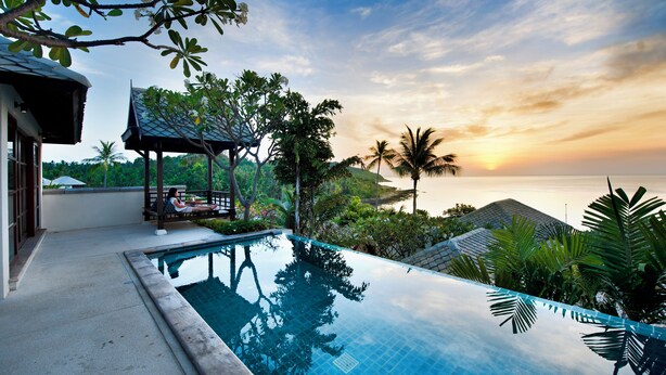 A swimming pool with a view of the sunset over the ocean, surrounded by lush greenery and palm trees. - Kanda Residences Pool Villas (Koh Samui)