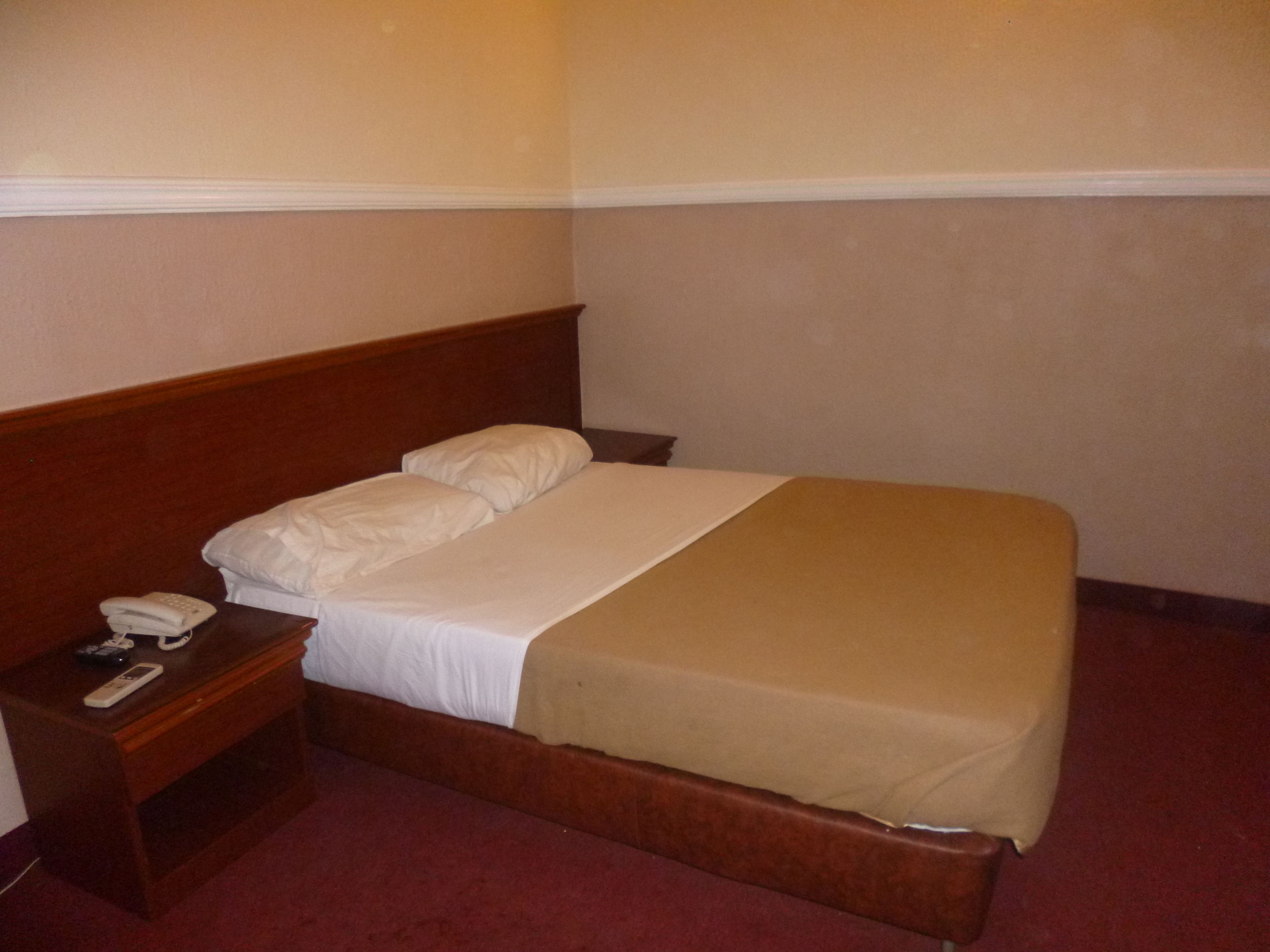 A single bed with a wooden headboard, a bedside table with a telephone and remote control, and a beige blanket.