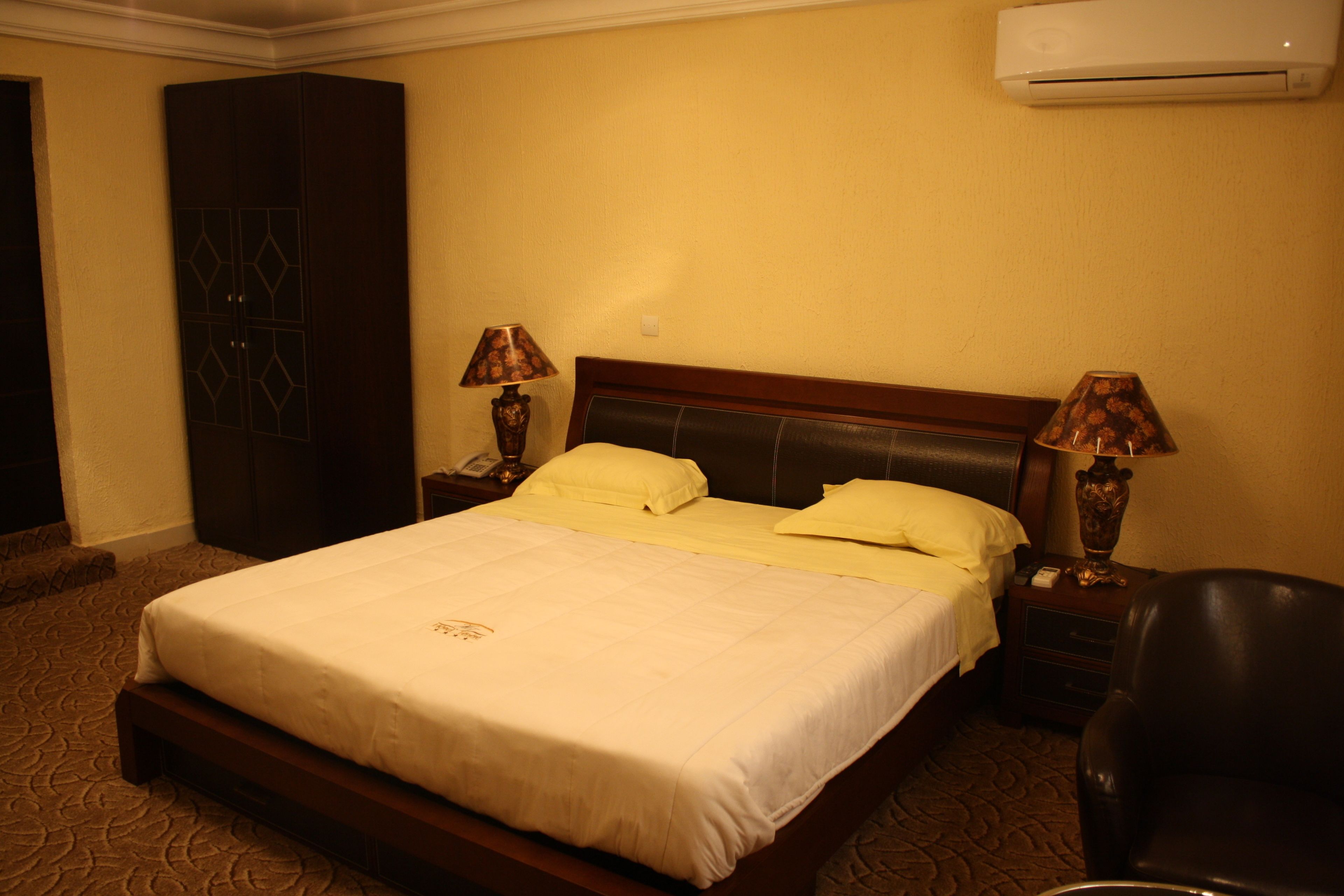 A hotel room with a large bed, two bedside tables with lamps, a wardrobe, and an air conditioning unit on the wall.