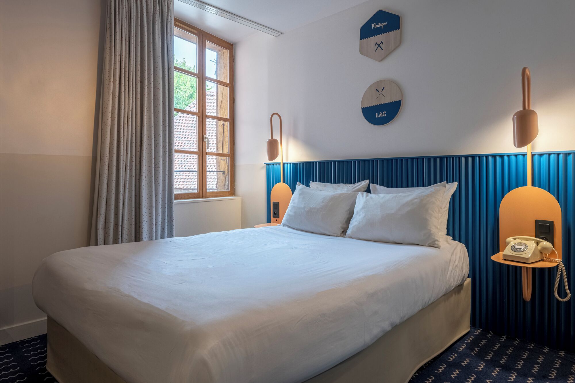Una habitación de hotel con una cama grande, una ventana con cortinas, un reloj de pared y un teléfono en la mesita de noche.