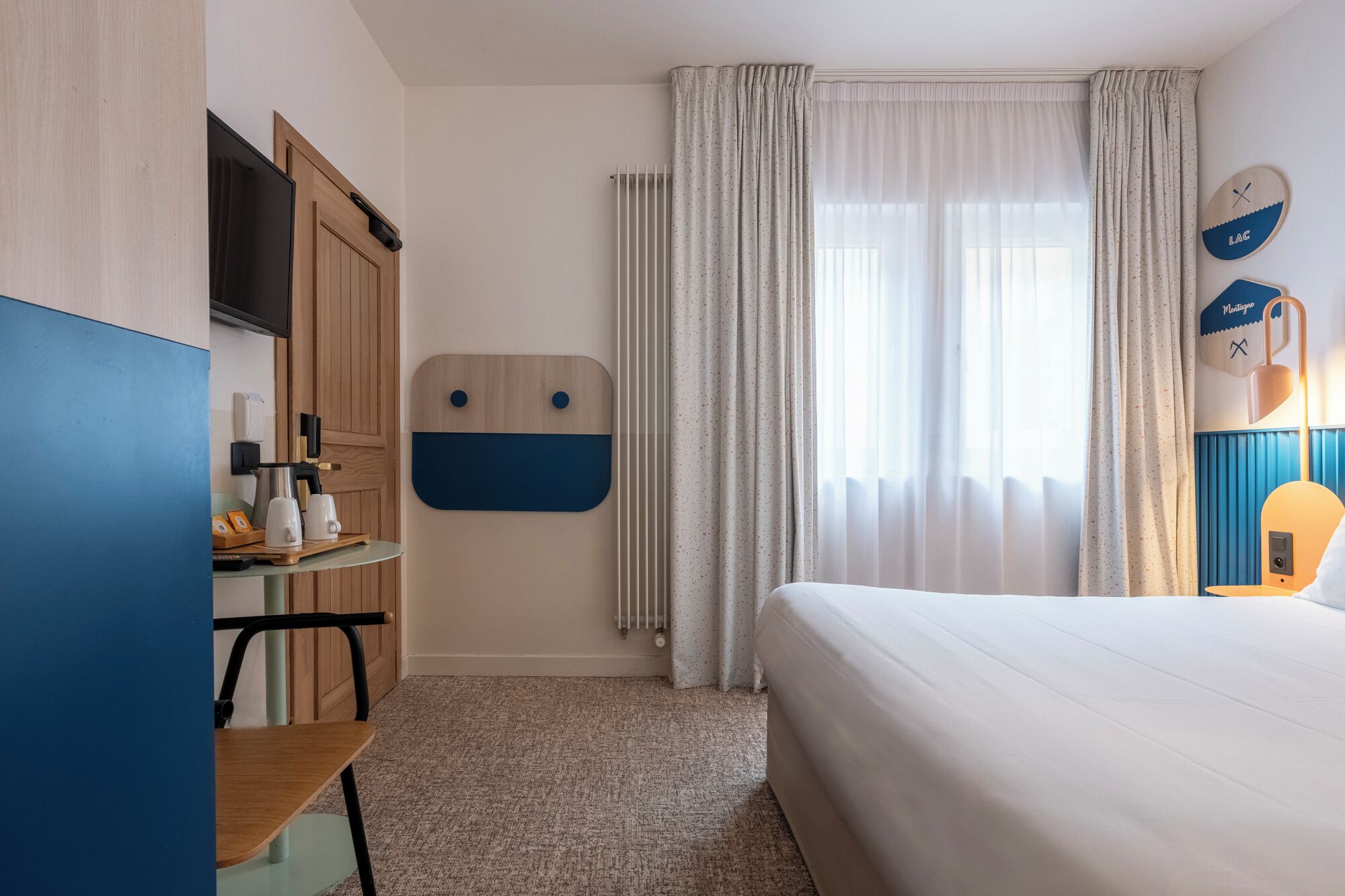 Quarto de hotel com cama, secretária com cadeira, radiador e cortinas.