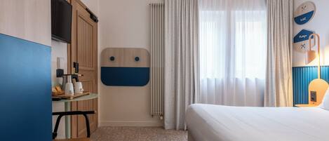 Quarto de hotel com cama, secretária com cadeira, radiador e cortinas.