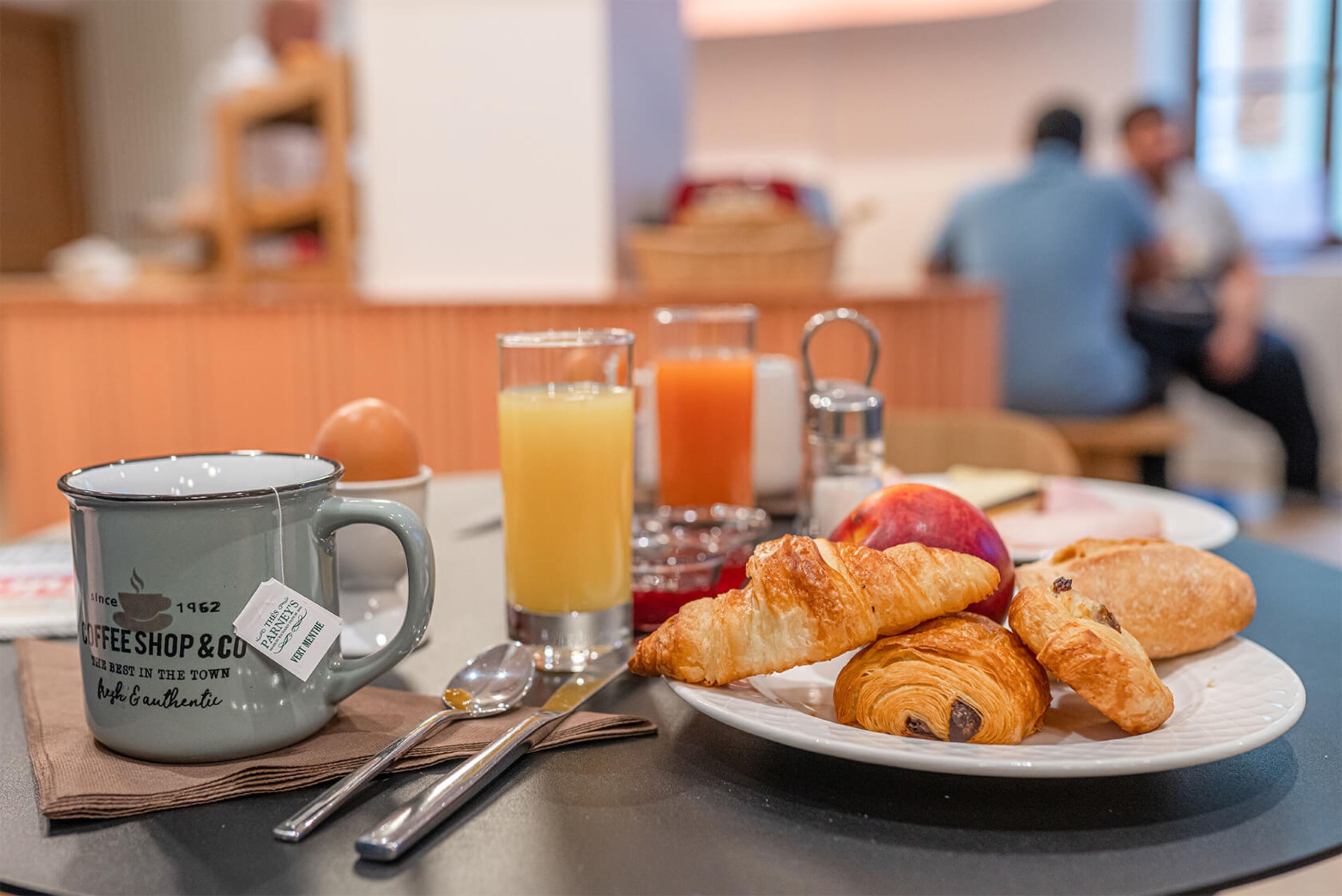A breakfast table with a coffee mug, croissants, a plate with an apple, and glasses of orange juice.