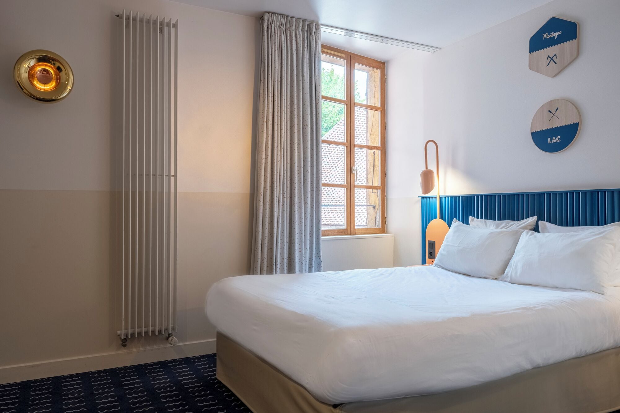 A hotel room with a large bed, a radiator, a window with curtains, and a wall clock.