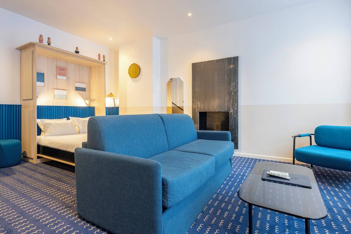 Une chambre d’hôtel moderne avec un canapé bleu, un lit avec des rideaux bleus, une petite table avec une télécommande et un téléviseur fixé au mur.