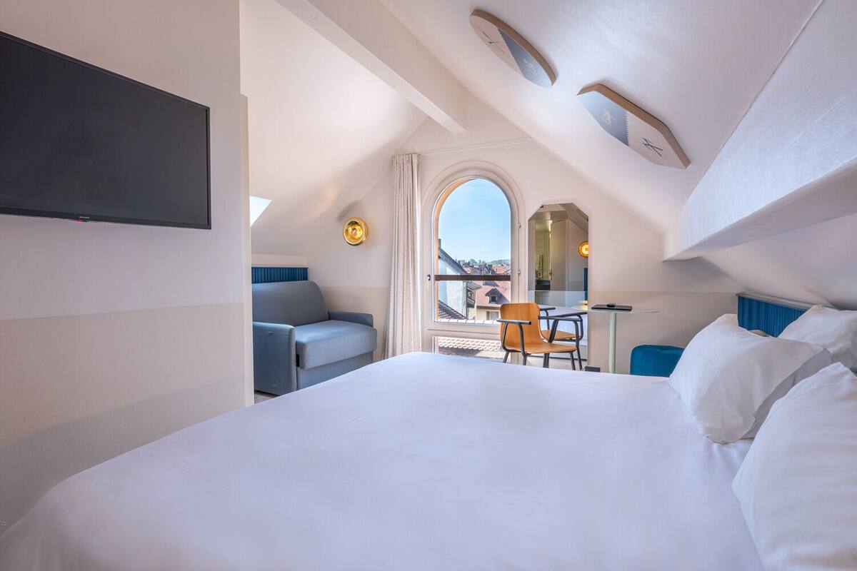 Une chambre d’hôtel moderne avec un grand lit, un bureau et une chaise. Un téléviseur est fixé au mur et une fenêtre offre une vue sur la ville.