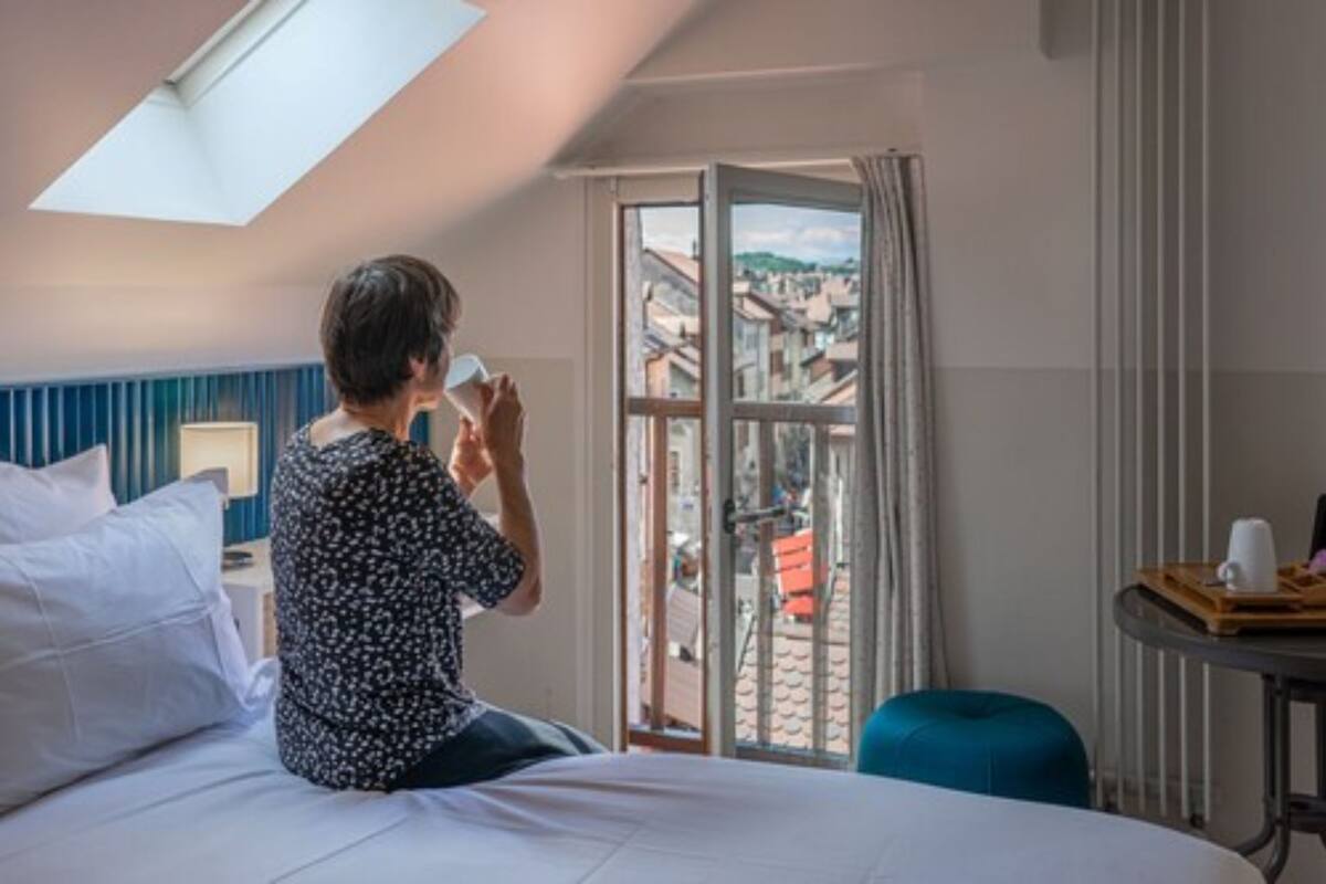 Une chambre d’hôtel avec un lit, une fenêtre donnant sur des immeubles, une table avec un plateau, et un pouf bleu.