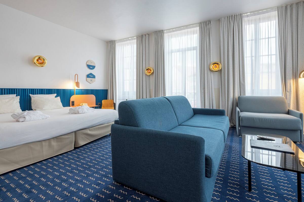 Une chambre d’hôtel comprenant un lit, deux fauteuils, un canapé, une petite table et une fenêtre avec des rideaux.