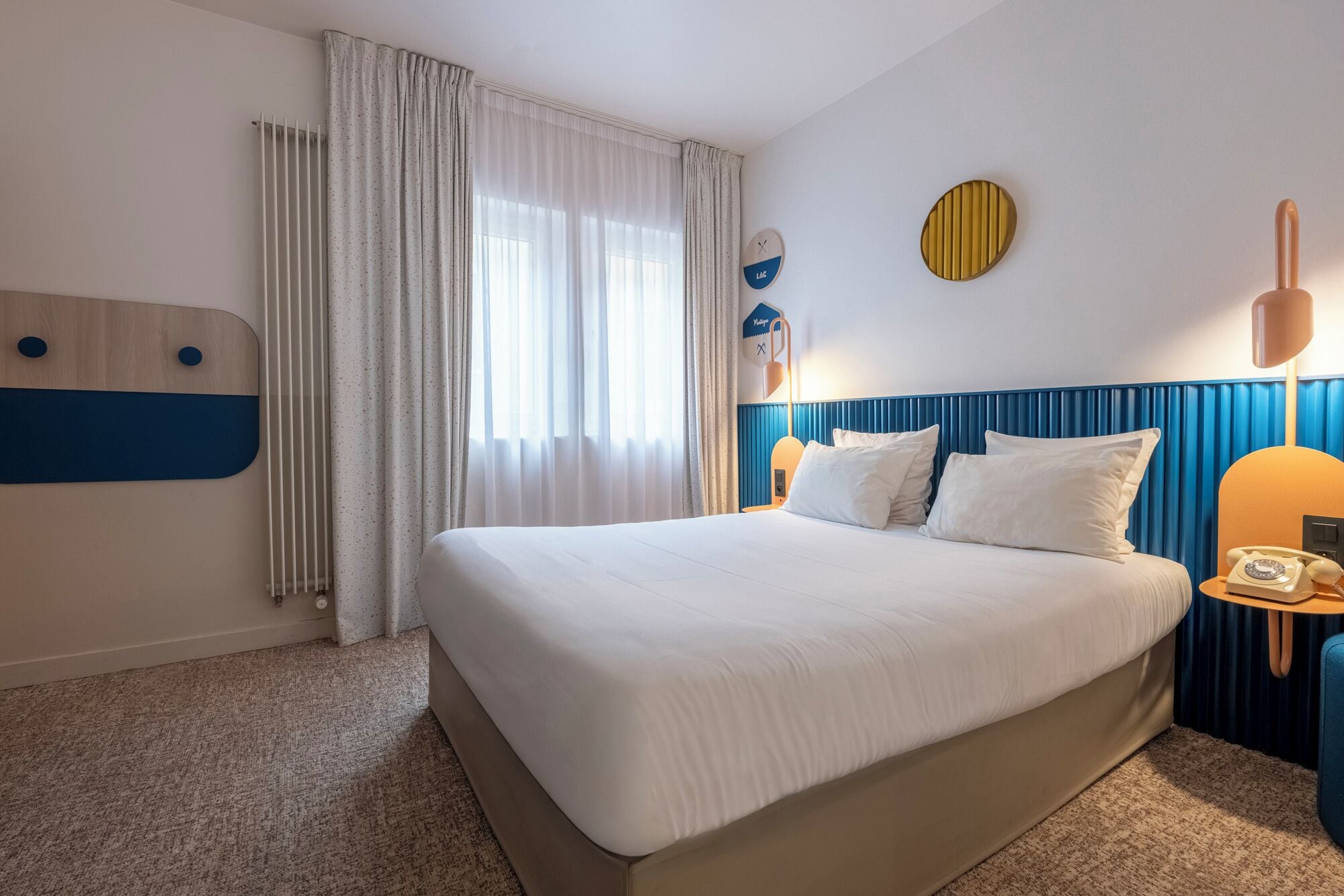 A hotel room with a large bed, a bedside table with a lamp, a wall-mounted phone, and a window with curtains.