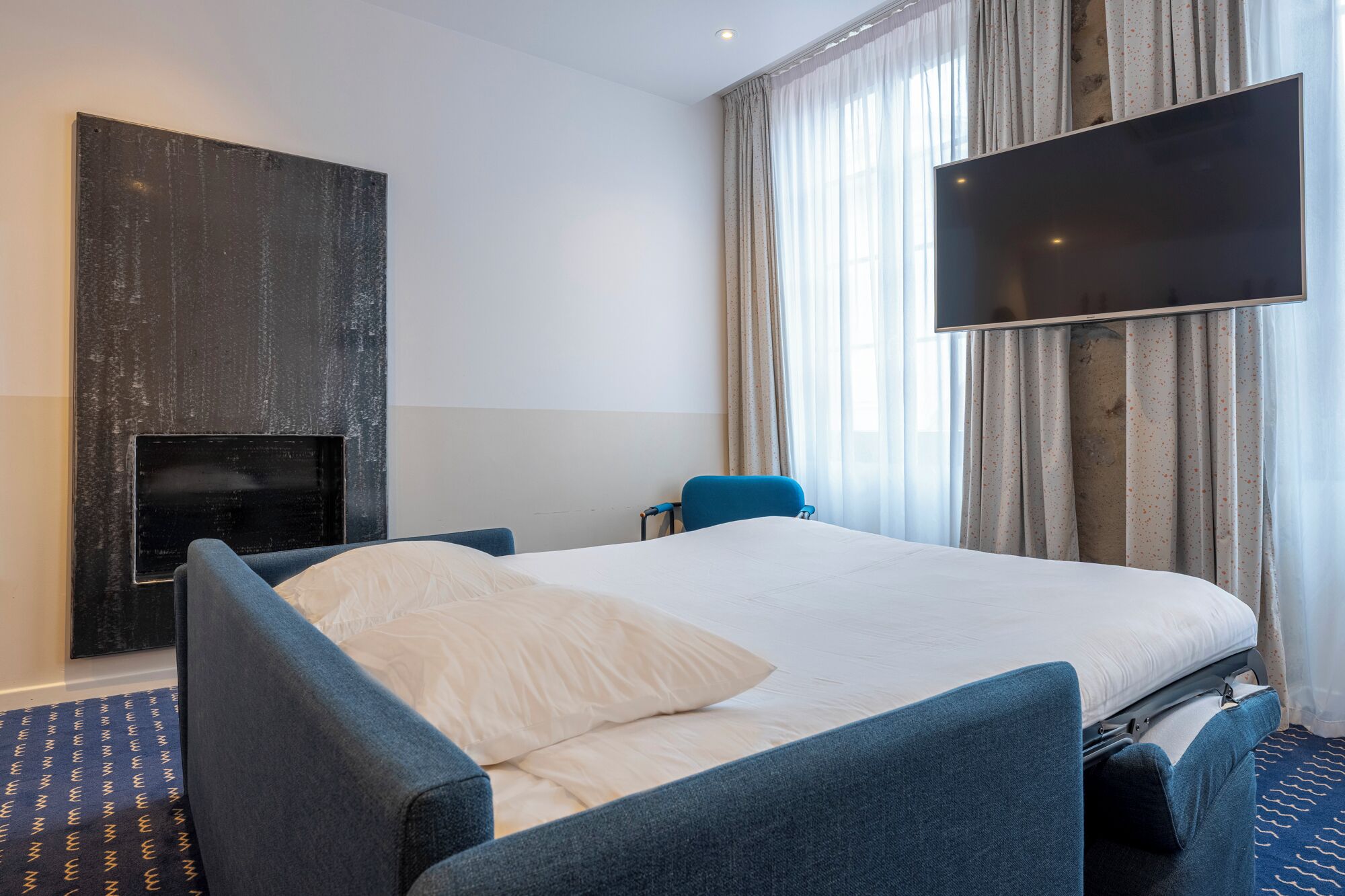 A hotel room with a bed, a chair, a flat-screen TV, and a fireplace.