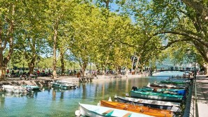 A serene riverside area with boats docked along the water's edge, surrounded by trees and a bridge in the background. - Hôtel du Palais de l'Isle (Annecy)