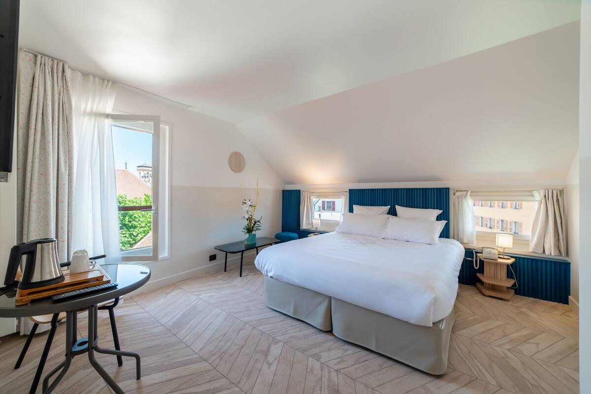 Une chambre moderne avec un grand lit, une petite table avec une bouilloire et une vue sur les bâtiments environnants.