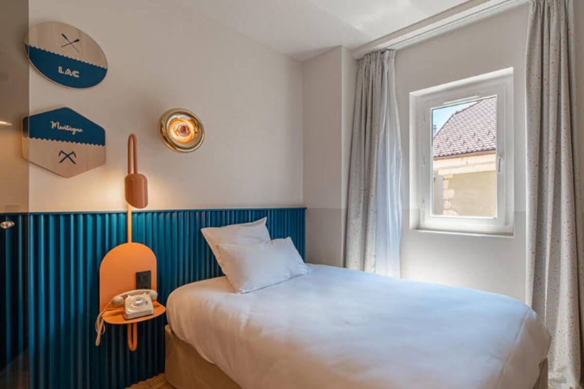 Une chambre d’hôtel avec un lit, une petite table avec un téléphone, une horloge murale et une fenêtre avec des rideaux.