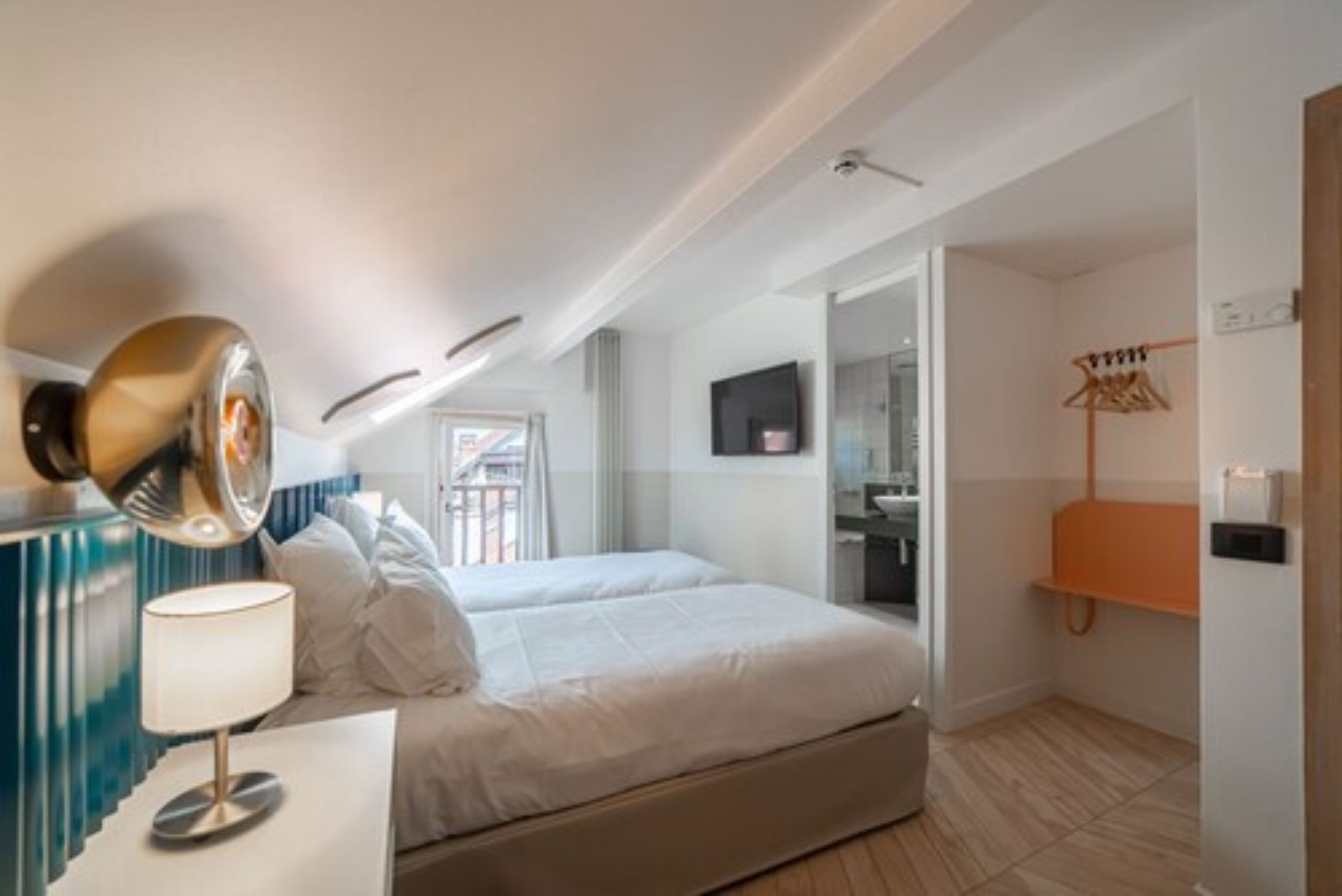 Une chambre d’hôtel avec un lit, une lampe de chevet, un téléviseur, un porte-manteau et une salle de bain visible à travers une porte ouverte.