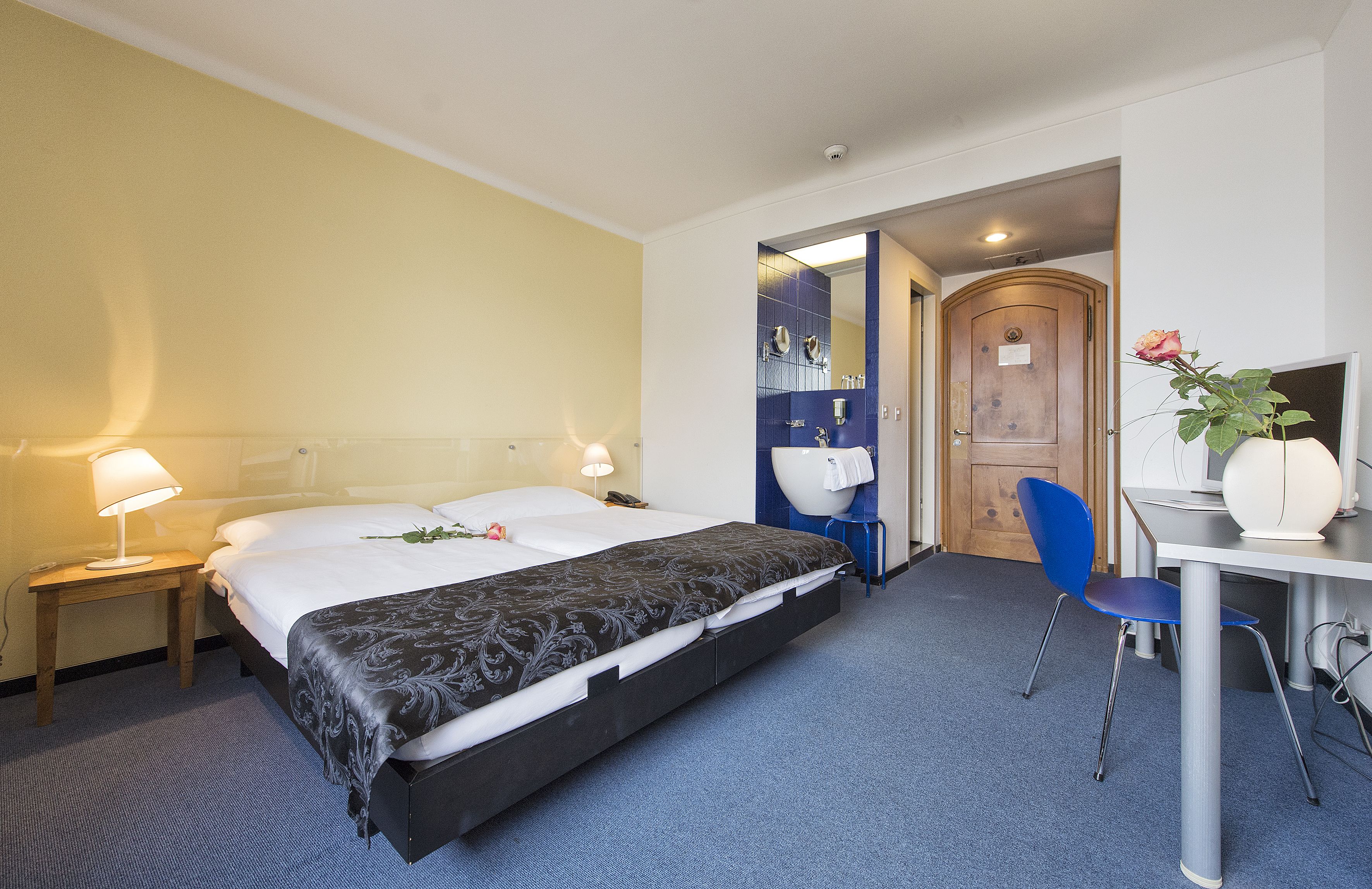 Ein Hotelzimmer mit Bett, Nachttischen, Schreibtisch, Stuhl und einem durchgegangenen Tür sichtbaren Badezimmer.