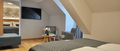Una habitación de hotel moderna con una cama grande, una zona de estar con dos sillones, una mesita con un jarrón y un amplio ventanal que proporciona luz natural.