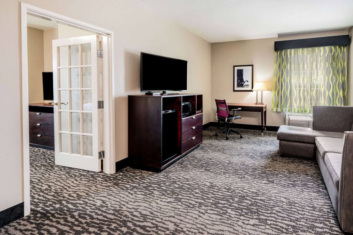 Ein Hotelzimmer mit Fernseher auf einem Standfuß, einem Schreibtisch mit Stuhl, einer Couch und einem gemusterten Teppich.