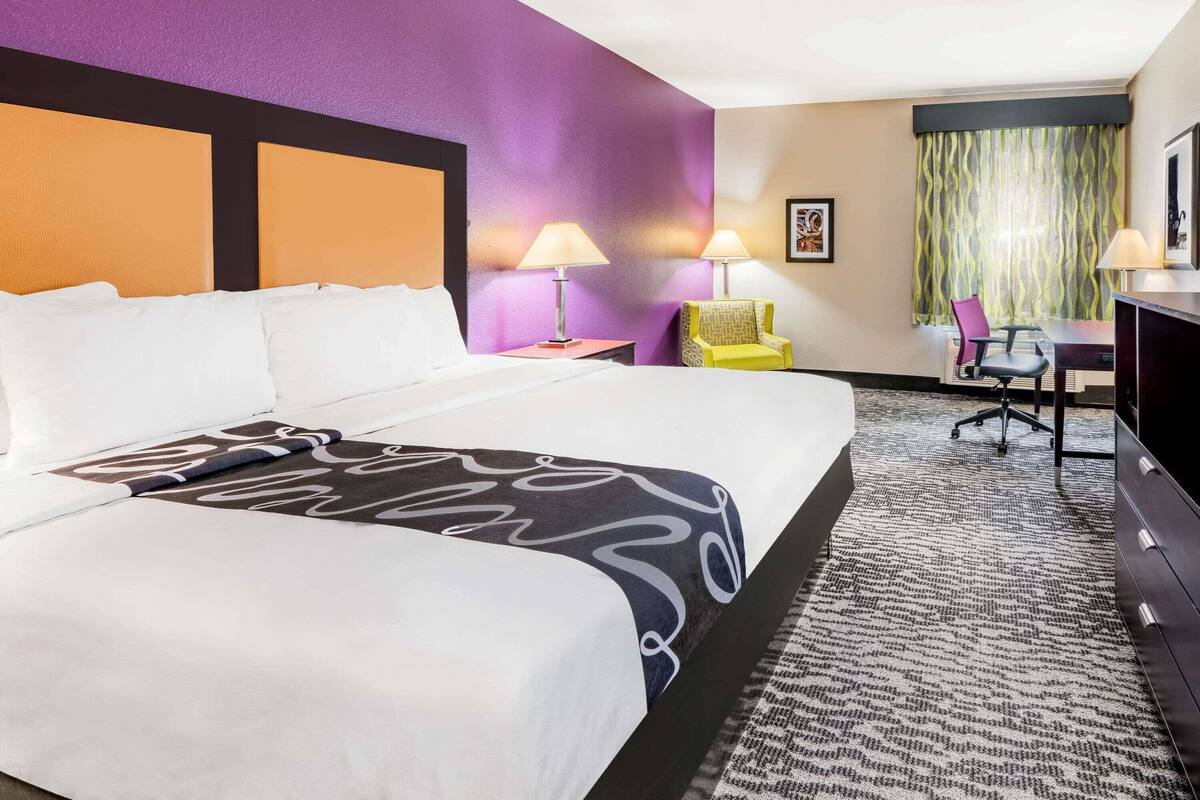 Ein Hotelzimmer mit einem großen Bett, zwei Nachttischlampen, einem Schreibtisch mit Stuhl und einem fensterbepflanzten Fenster.