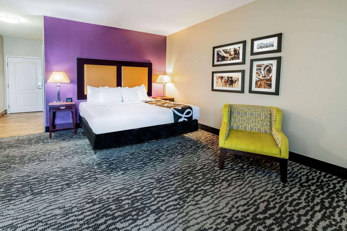 Ein Hotelzimmer mit einem Bett, einem Sessel, einem Nachttisch, einer Lampe und gerahmten Bildern an der Wand.
