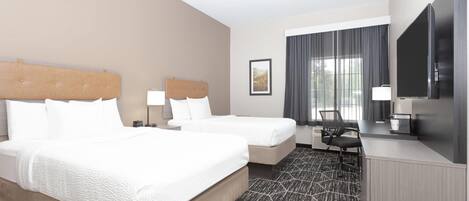 A hotel room with two beds, a desk, a TV, and a large window with curtains.