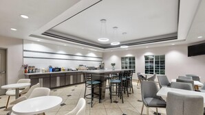 Un área de comedor de hotel moderna con mesas redondas, taburetes de bar y una cocina al fondo.
