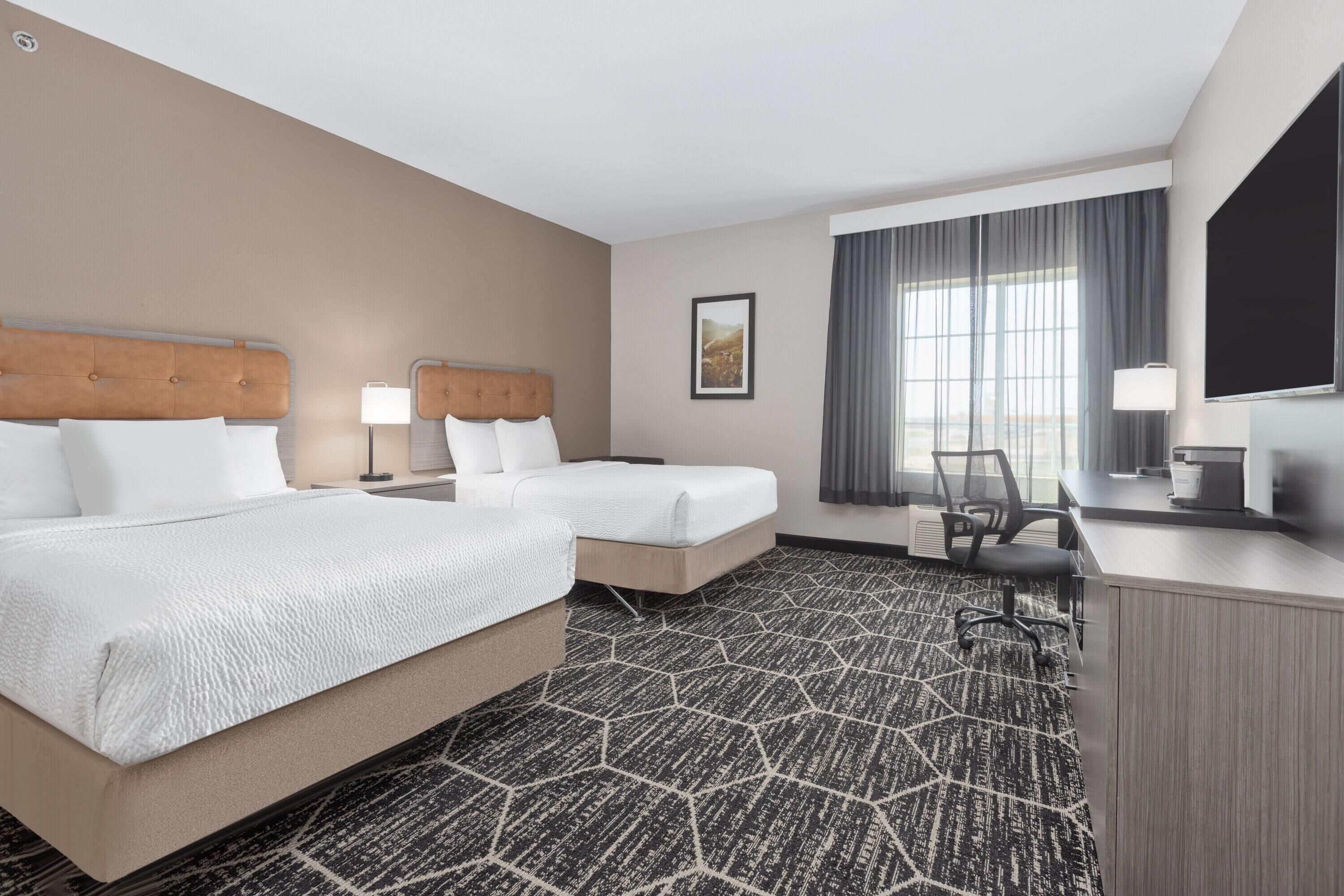 A hotel room with two beds, a desk, a chair, a TV, and a large window with curtains.