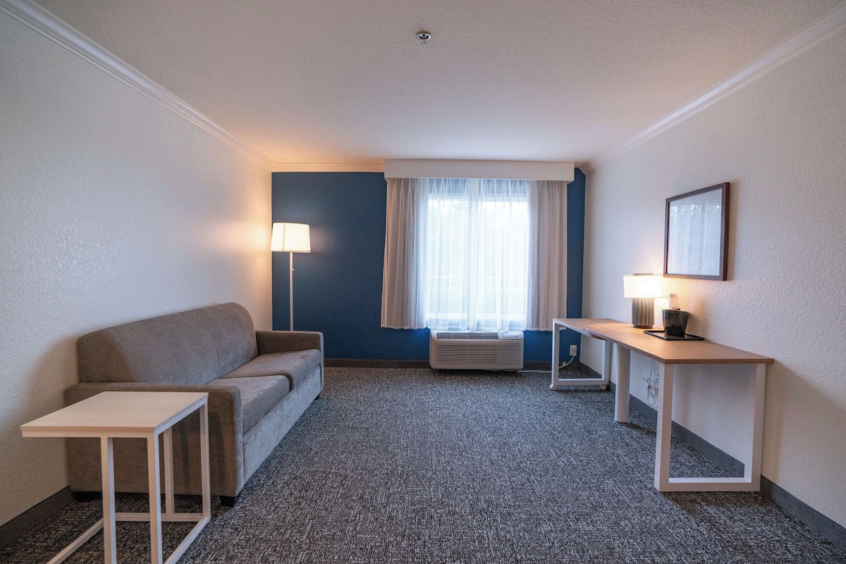 Una habitación de hotel con sofá, una mesita, una lámpara y una ventana con cortinas.