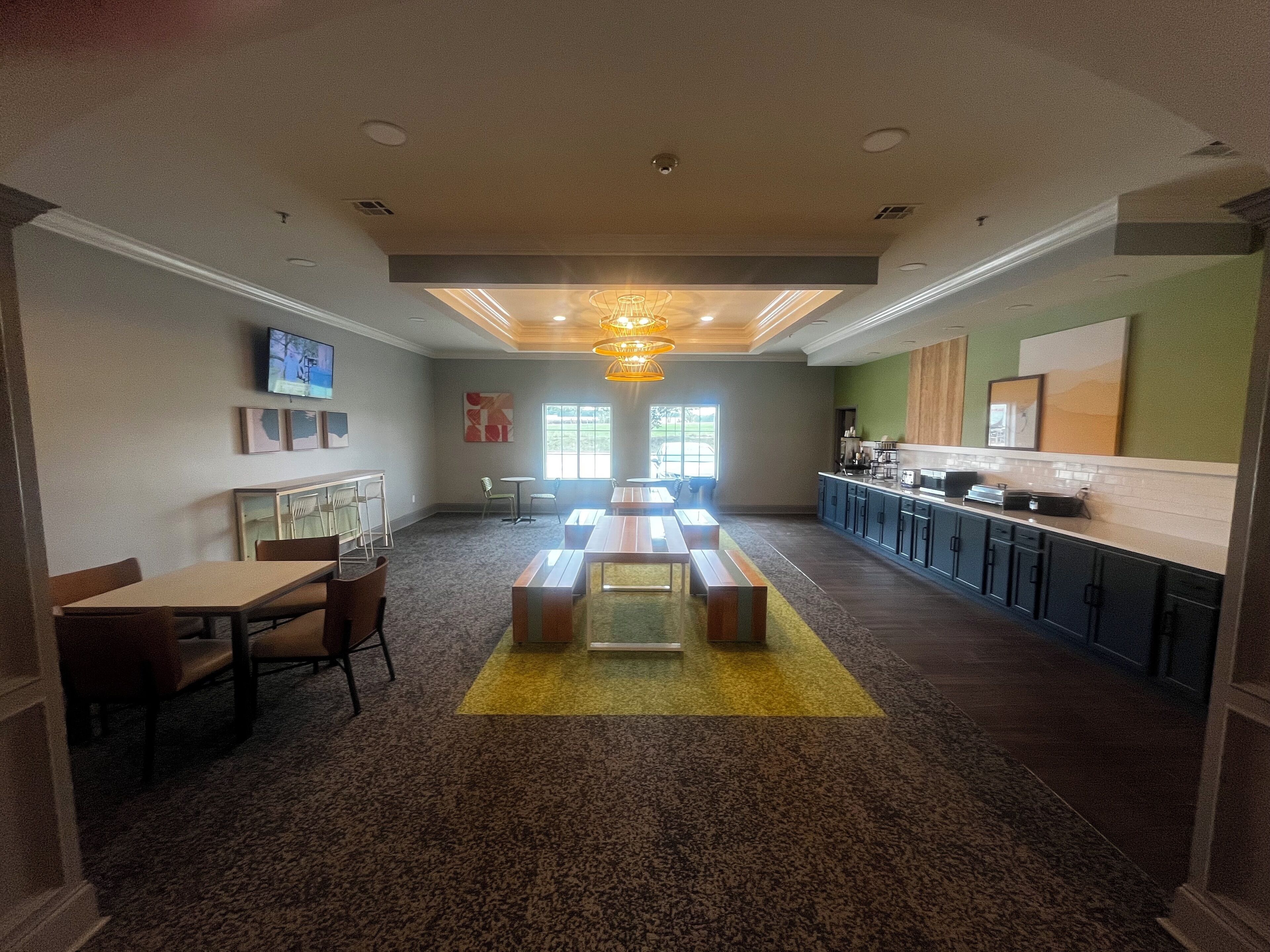 A spacious room with a long table and benches, a kitchen area with a coffee machine, and a television on the wall.