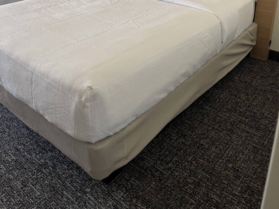 Habitación de hotel con dos camas, una cama grande con cabecera color beige y una cama más pequeña con cabecera de madera. Hay dos mesitas de noche con lámparas, un televisor montado en la pared y cuadros enmarcados sobre las camas.