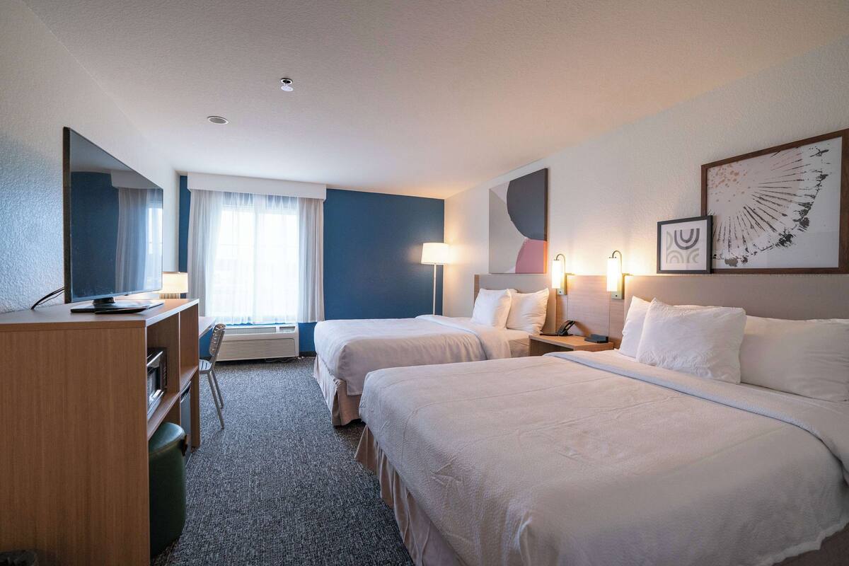 Habitación de hotel con dos camas, un escritorio, una silla, una lámpara y un ventanal con cortinas.