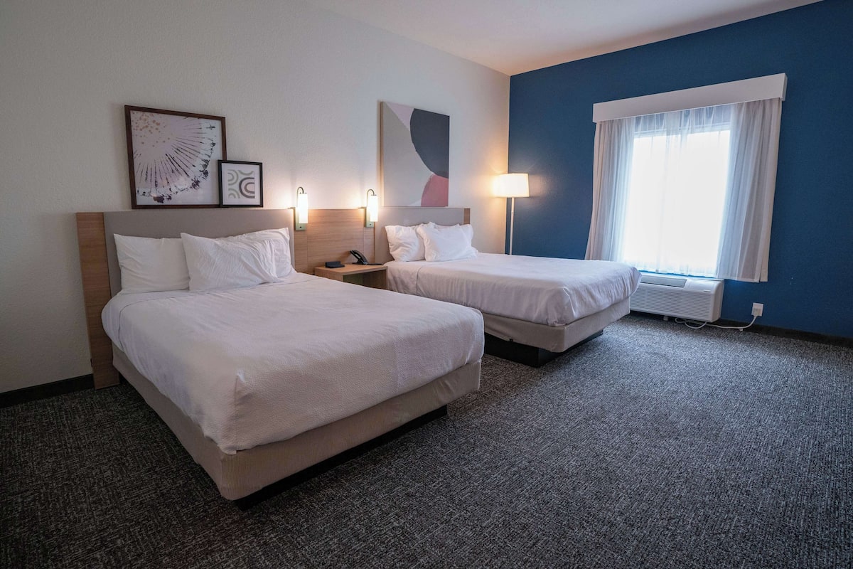 Habitación de hotel con dos camas, una mesita de noche con lámpara, una obra de arte en la pared y una ventana con cortinas.