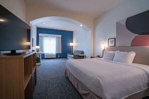 Habitación de hotel con una cama grande, un televisor sobre un escritorio de madera, una silla, una lámpara y una ventana con cortinas.