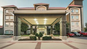 A hotel entrance with a covered walkway, columns, and a landscaped area with shrubs. - La Quinta Inn & Suites by Wyndham San Antonio Northwest (San Antonio)