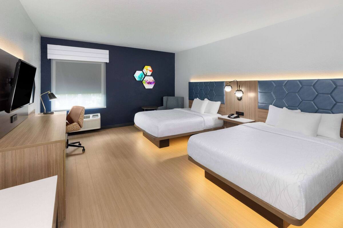 Une chambre d’hôtel avec deux lits, un bureau, une chaise, une télévision et une fenêtre avec des stores.