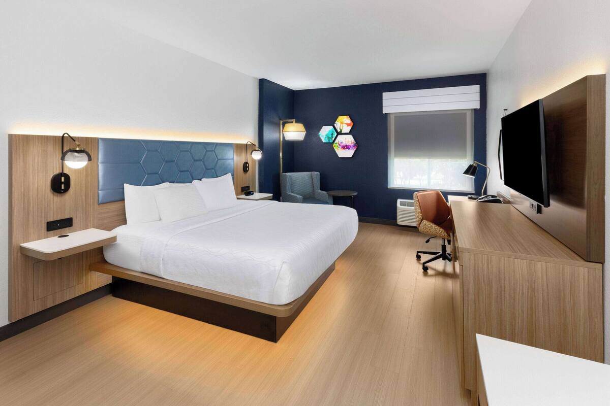 Une chambre d’hôtel moderne dotée d’un grand lit, de tables de chevet, d’un bureau, d’une chaise, d’un téléviseur à écran plat et d’une fenêtre avec des stores.