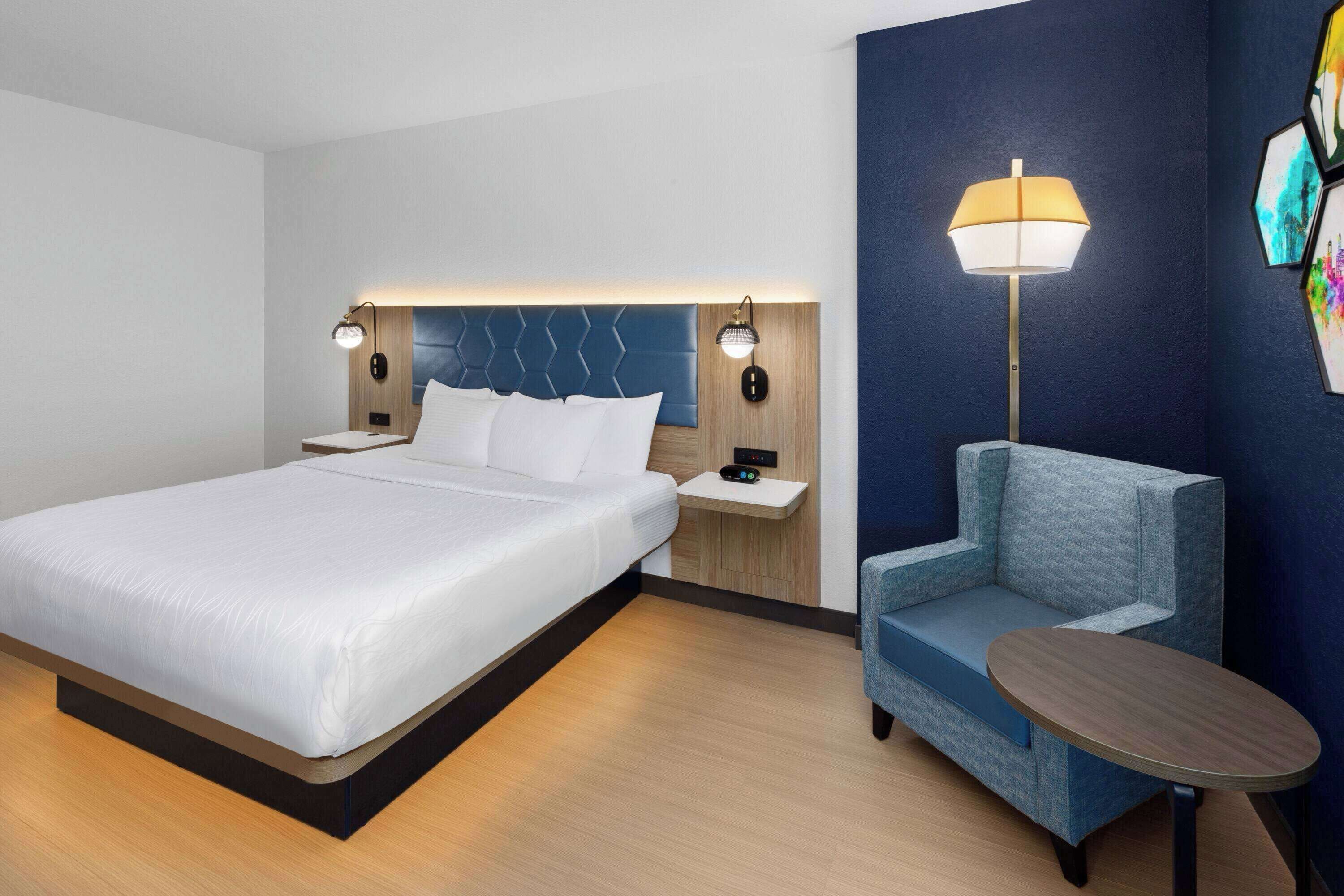 Una habitación de hotel moderna con una cama grande, una mesita de noche, un sillón azul y una pequeña mesa redonda.
