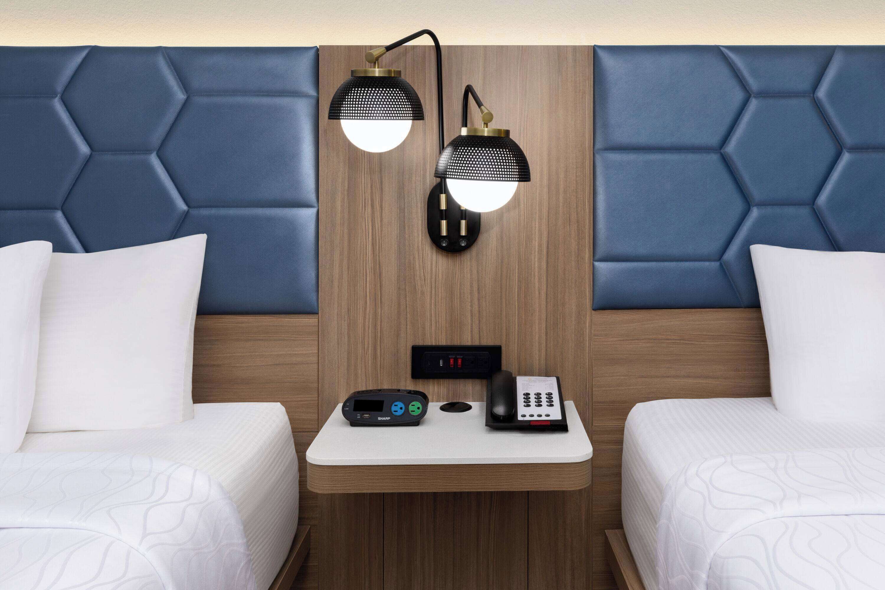 A hotel room with a wooden headboard, two beds, a small bedside table with a phone and a remote control, and two wall-mounted lamps.