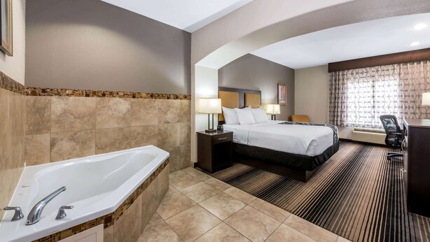 A hotel room with a large bathtub, a bed with a headboard, a desk with a chair, and a window with curtains.