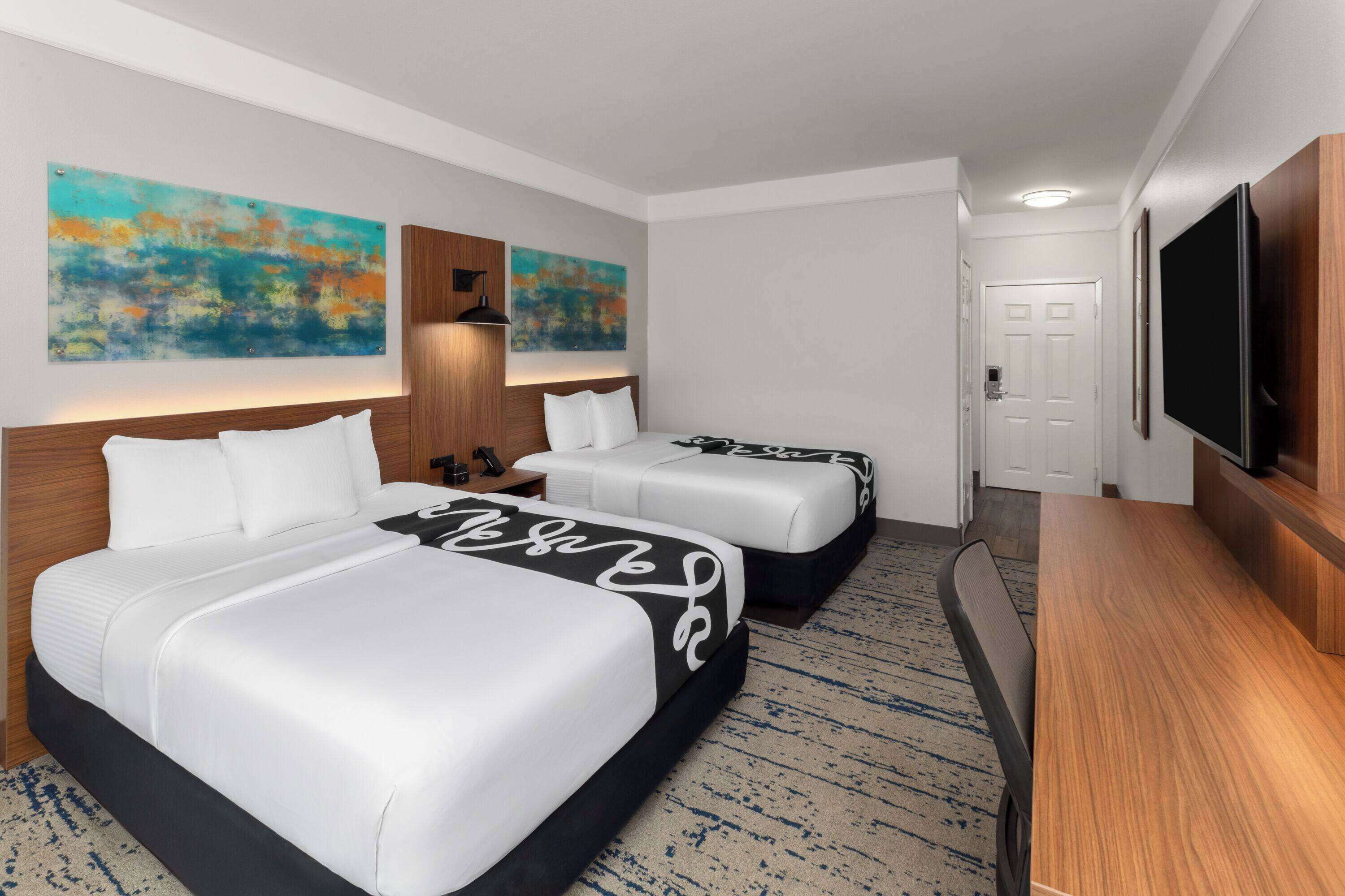 A hotel room with two beds, a flat-screen TV, a desk, and artwork on the walls.