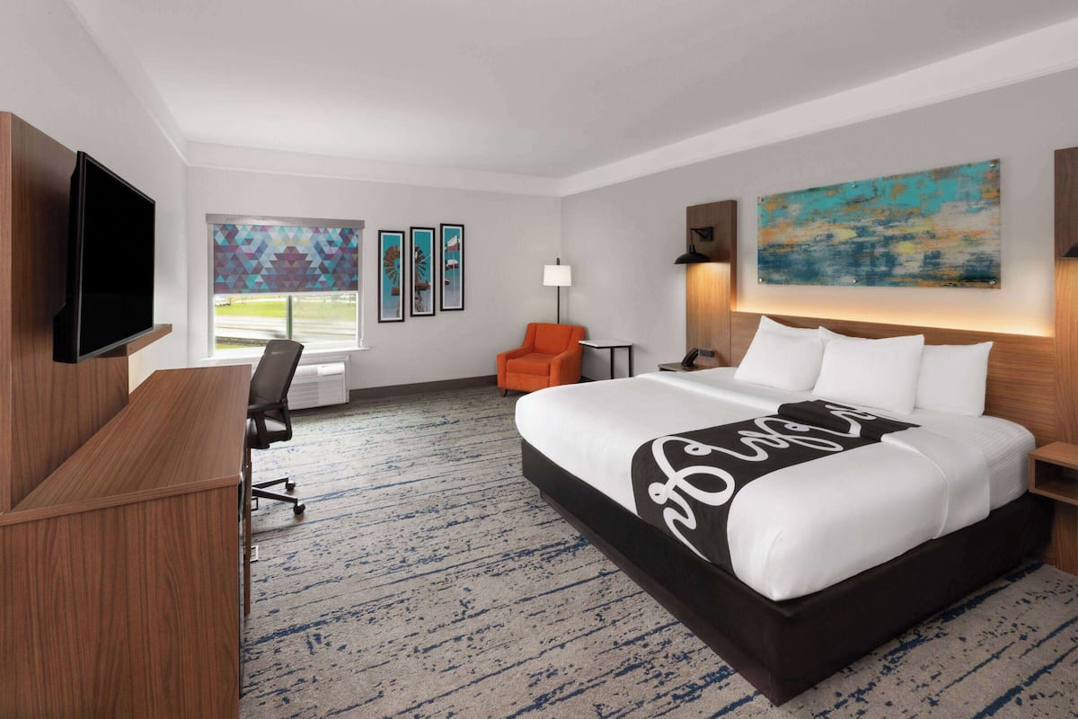 A hotel room with a large bed, a desk, a TV, an orange chair, and artwork on the walls.