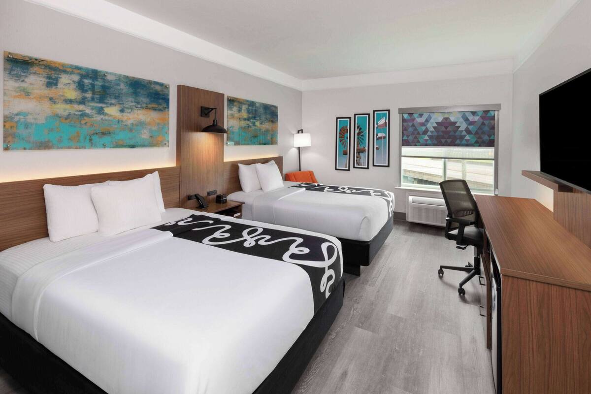 A hotel room with two beds, a desk, a TV, and artwork on the walls.