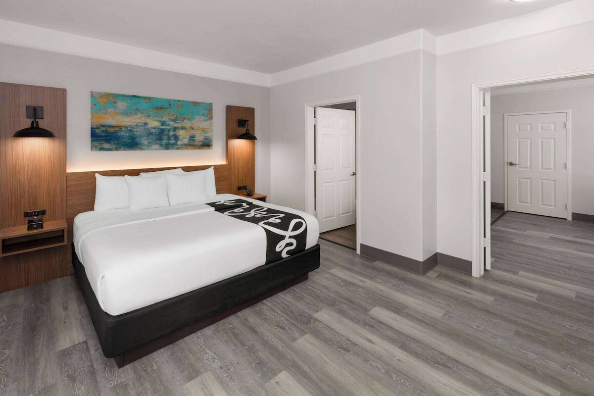 A modern bedroom with a large bed, wooden headboard, two bedside lamps, a painting on the wall, and a door leading to another room.