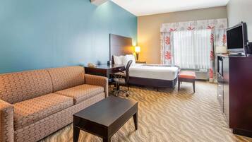 A hotel room with a sofa, a desk, a bed, a TV, and a window with curtains.
