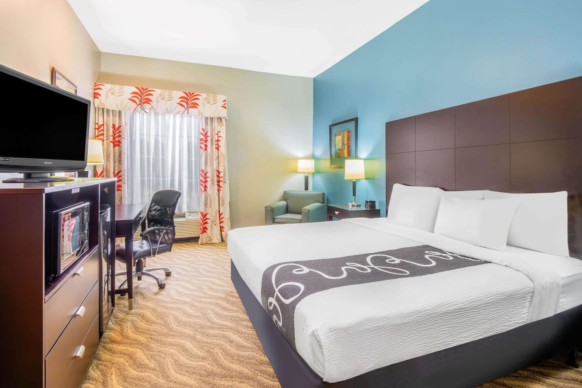 Une chambre d’hôtel avec un lit, un bureau avec une télévision, une chaise, une lampe et une fenêtre avec des rideaux.