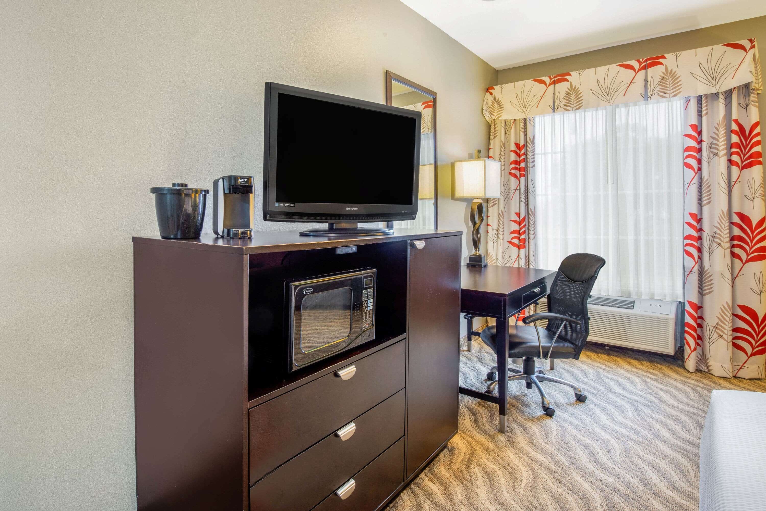 A hotel room with a flat-screen TV, a microwave, a desk with a chair, and a window with curtains.