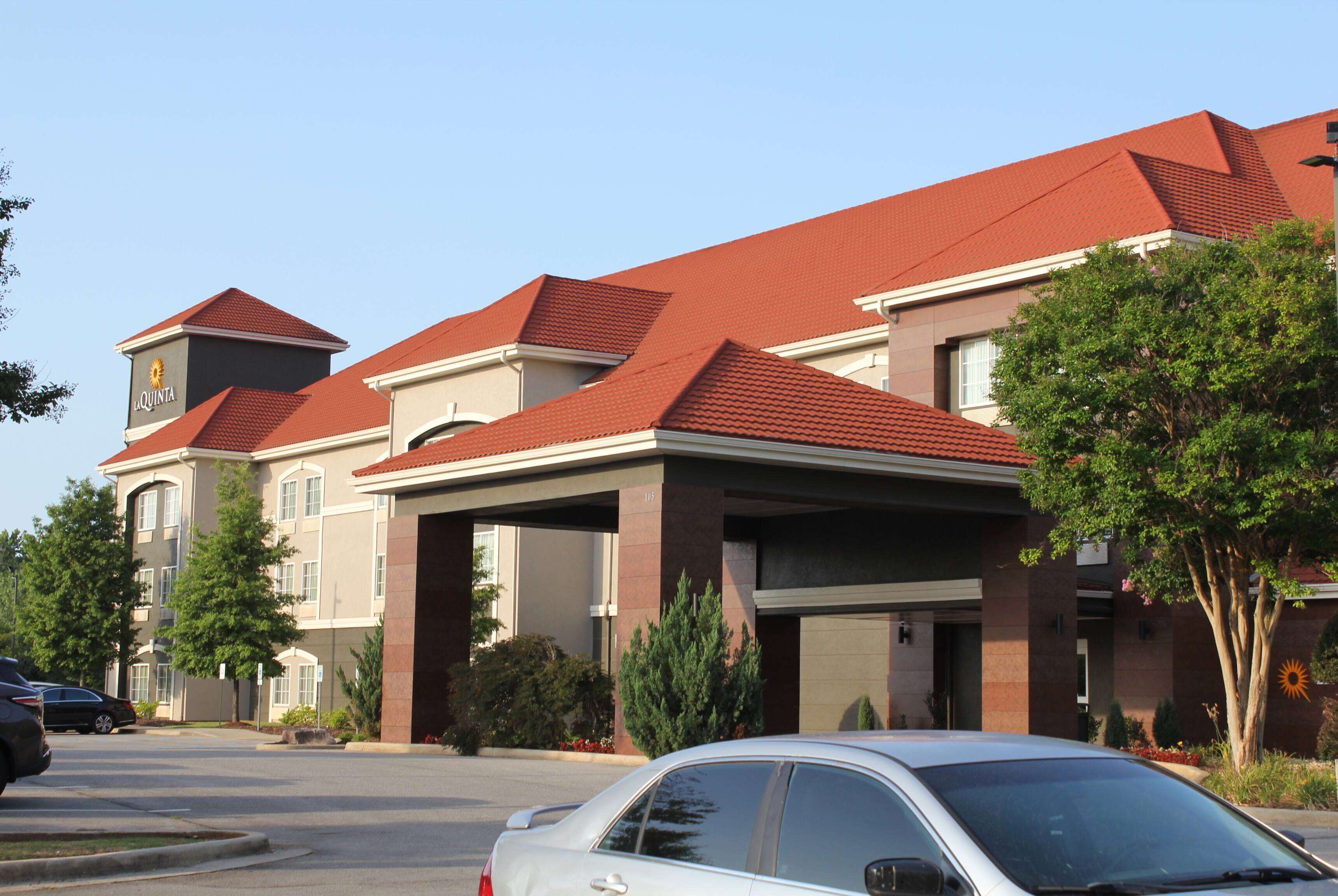 A multi-story hotel with a red roof, a covered parking area, and a well-maintained landscape with trees and shrubs.